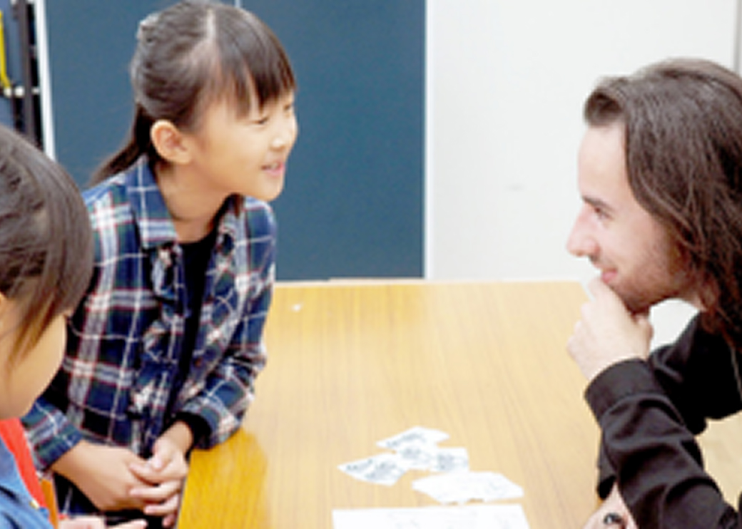 外国人講師と子どもがテーブルを挟んで向かい合い、笑顔で話している様子。学びや交流をテーマにした活動シーン