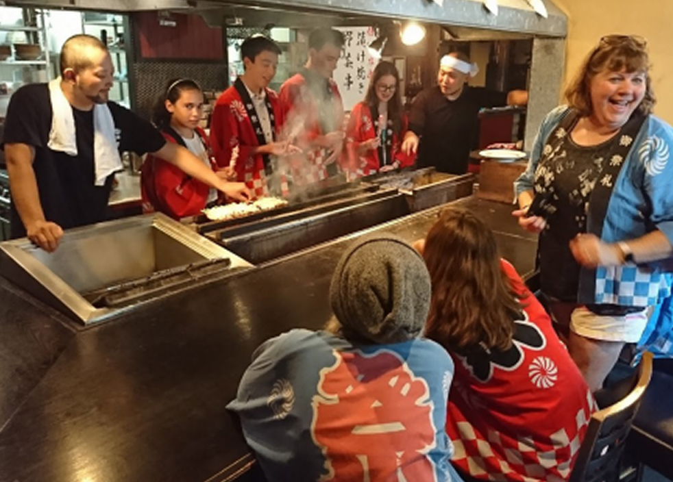 外国人学生が鉄板焼きの店で料理を体験しながら日本人スタッフと交流している様子