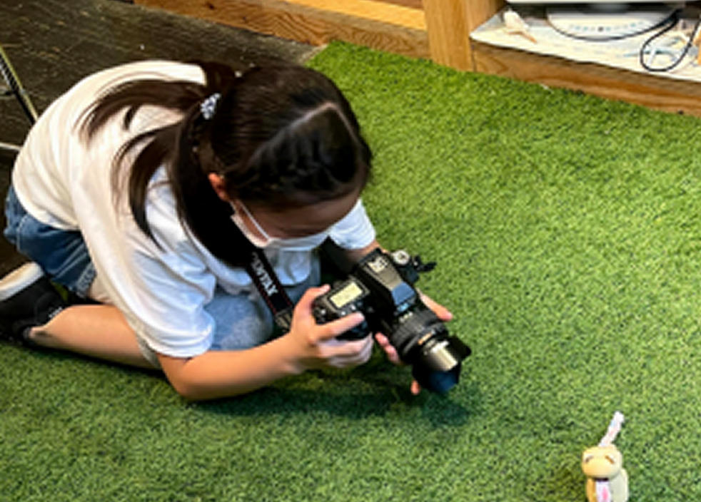芝生の上でカメラを構え、小さなぬいぐるみを撮影している女の子の様子。取材活動の一環として写真を撮っている