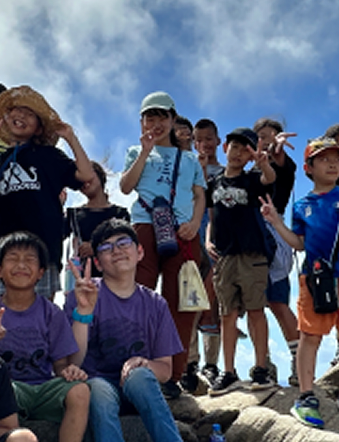 青空の下で子どもたちが集まり、笑顔でピースサインをしている様子。登山や野外活動に参加しているグループの集合写真
