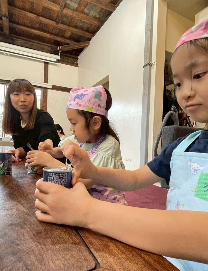 エプロンと三角巾をつけた子どもたちが、大人と一緒にテーブルで作業をしている様子