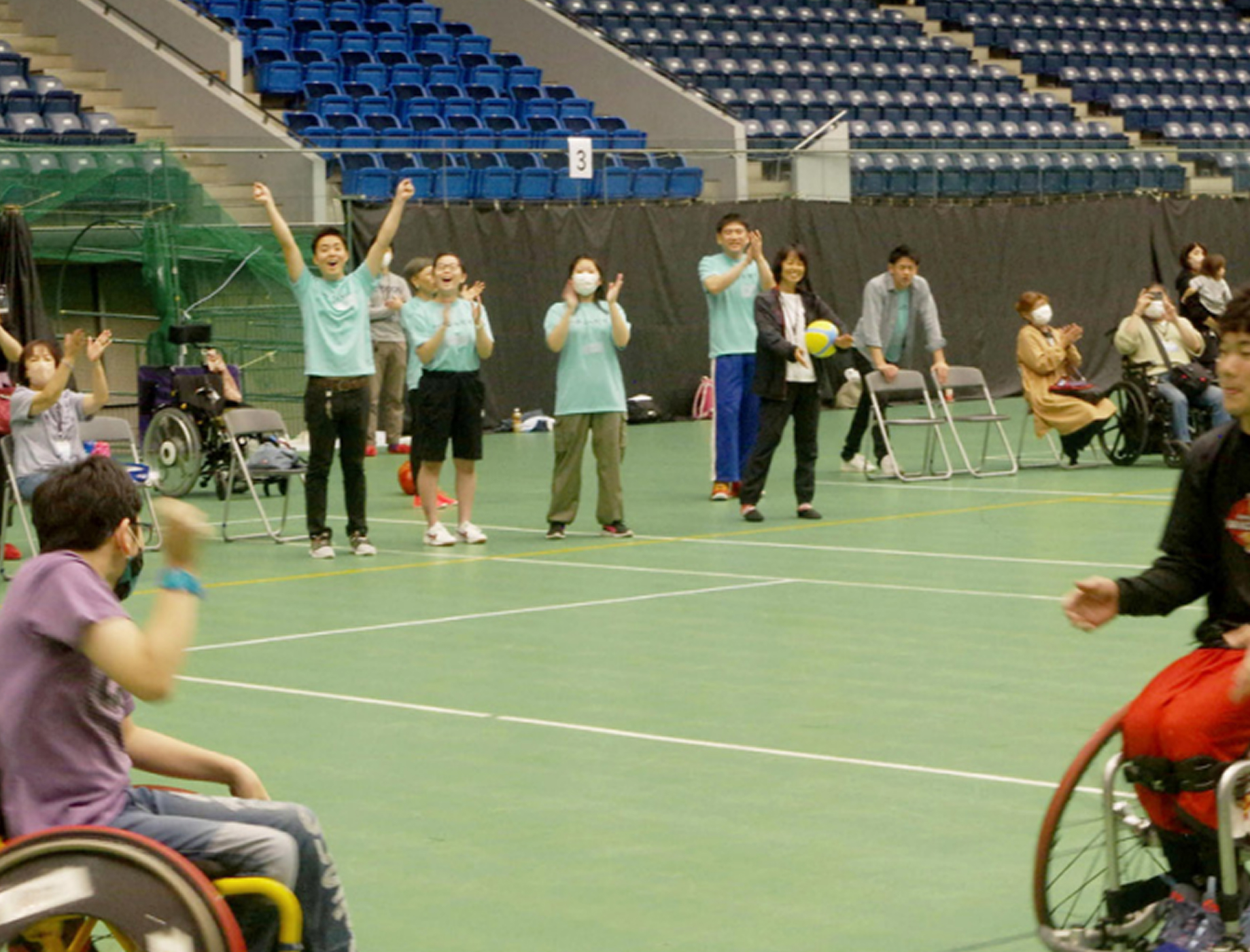 体育館で車いすの参加者とサポートスタッフが一緒に運動を楽しんでいる様子。みんなで手を挙げて盛り上がっている