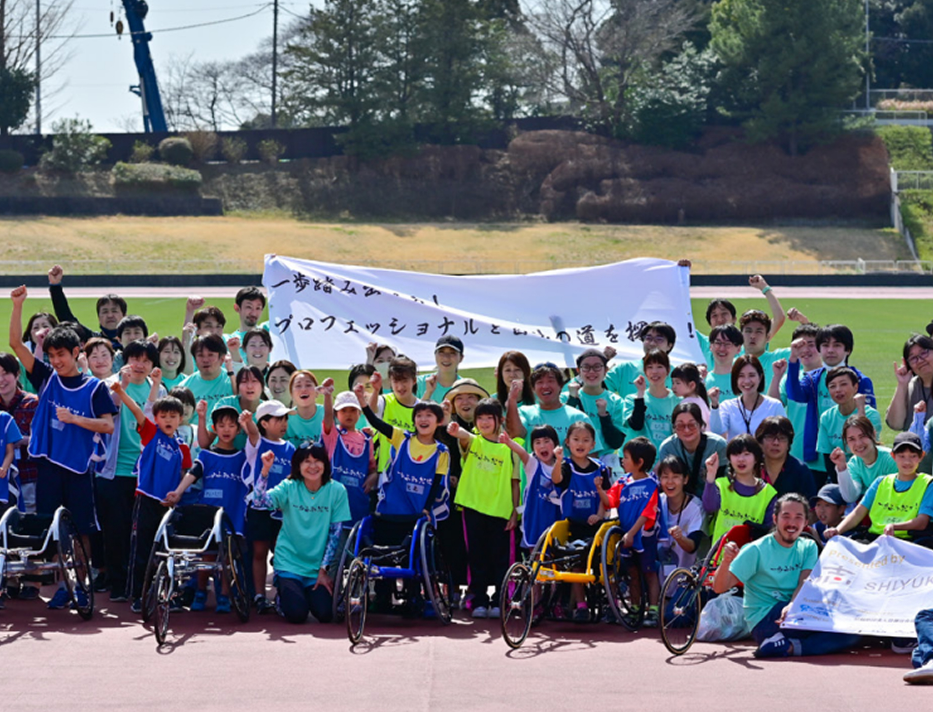 屋外の運動場で子どもたちと大人が集合写真を撮っている様子。車いすの参加者も含めて全員が笑顔でカメラに向かっている