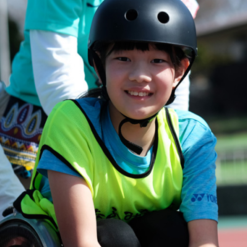 ヘルメットと蛍光ベストを着けたこどもが笑顔でカメラを見ている様子。屋外でスポーツに挑戦している