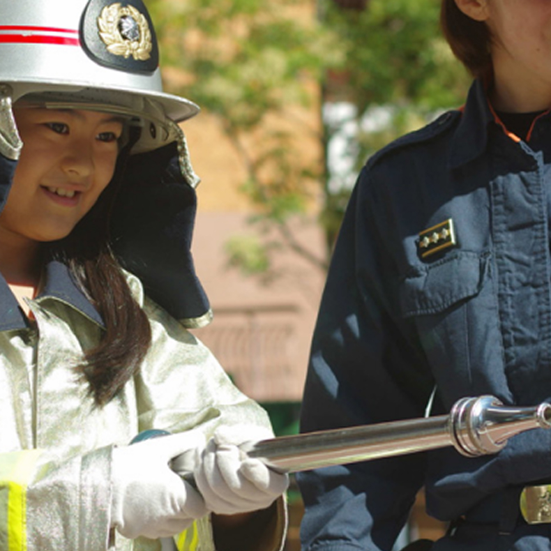 消防士の制服を着た子どもが放水用ホースを持ち、消防士と一緒に体験している様子