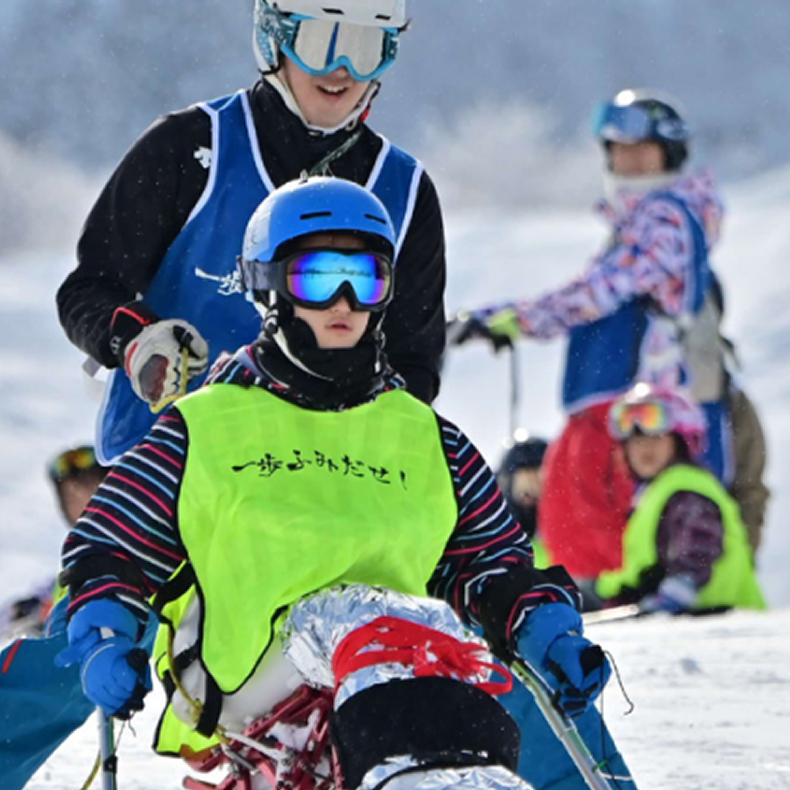 雪の上でヘルメットとゴーグルをつけた人が座ってスキーを楽しんでいる様子。後ろにはサポートスタッフの姿もある