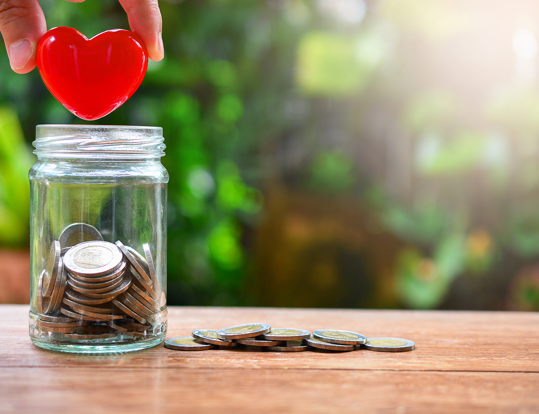 木のテーブルの上に置かれた瓶にコインが入っており、その上に赤いハート型のオブジェを持った手がかざされている。寄付や支援を象徴するイメージ写真