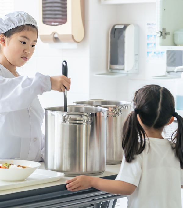 コック帽をかぶった子どもが大きな鍋をかき混ぜて料理をしている様子