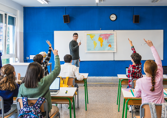 教室で子どもたちが手を挙げて先生の話を聞いている様子。学びと参加をテーマにした授業風景